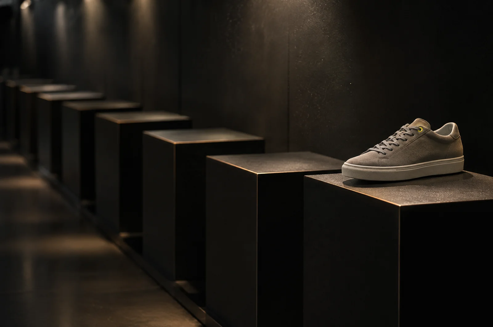 Eine Reihe leerer dunkler Präsentationssockel in einem Laden, nur auf dem vordersten steht noch ein einzelner grauer Wildleder-Sneaker mit gelber Öse.