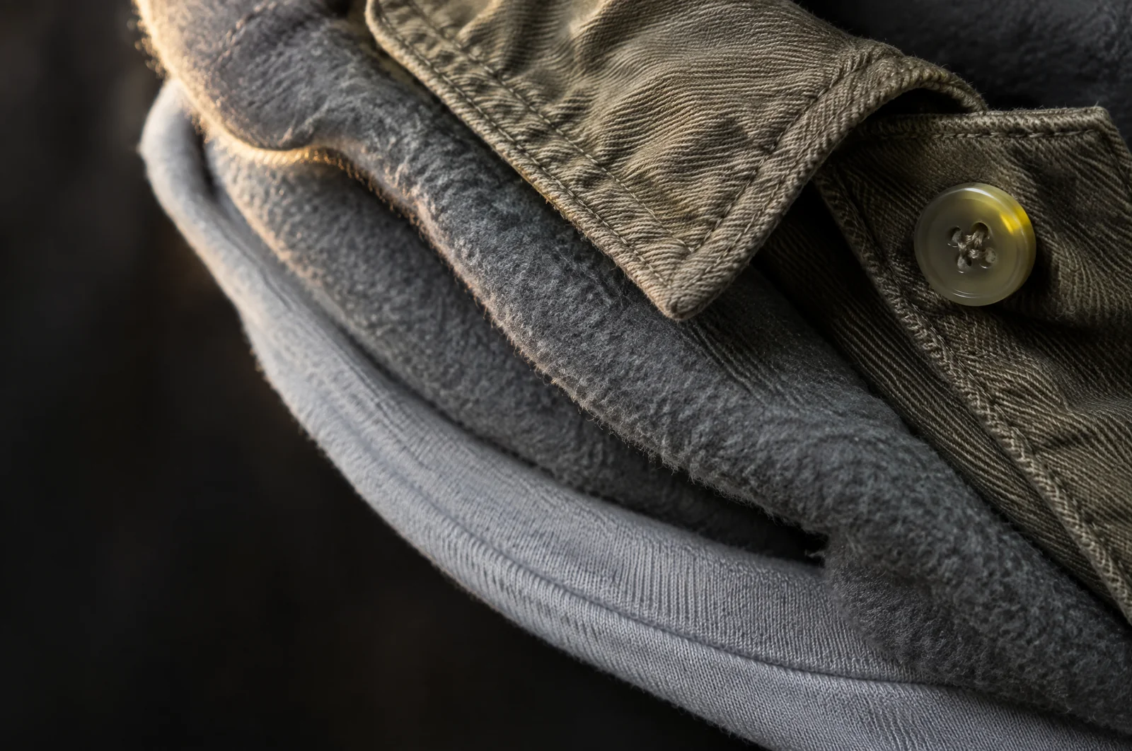 Drei Stofflagen übereinander am Kragen: unten ein dünnes Jersey, darüber die weiche Kapuzenkante eines Hoodies, oben der schwere Kragen mit Knopfleiste.