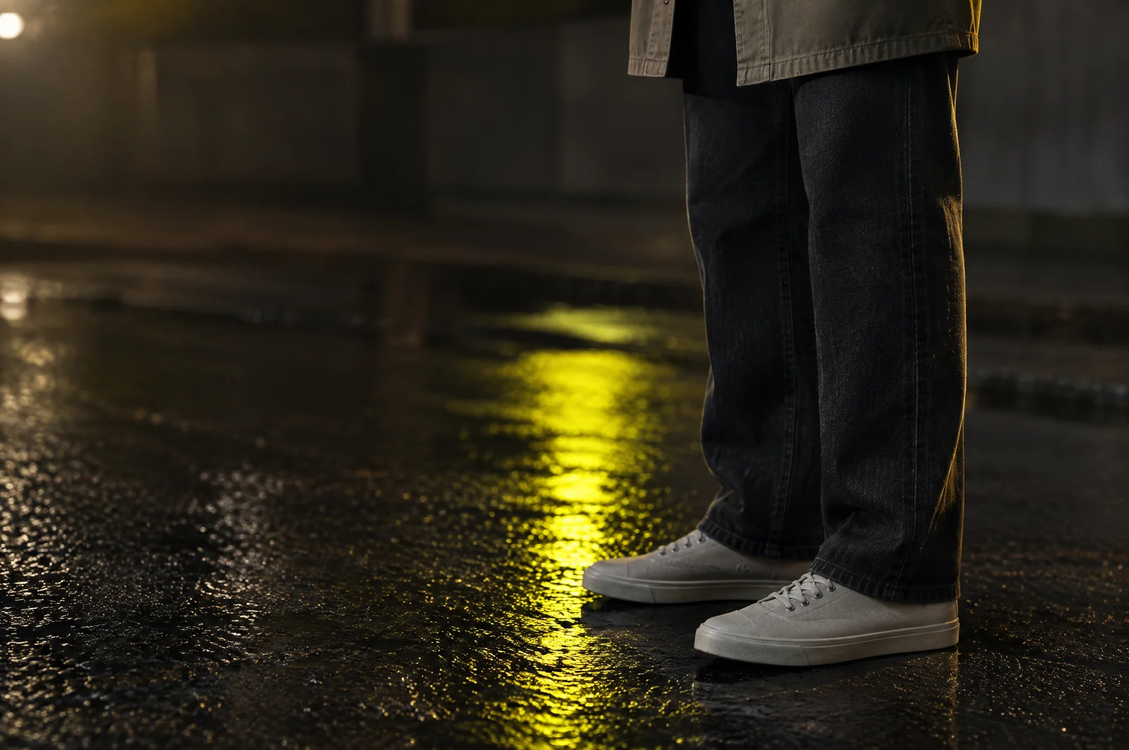 Eine dunkle gerade Jeans über flachen hellen Sneakern auf nassem Asphalt, darüber endet der Saum eines offenen Overshirts an der Hüfte.