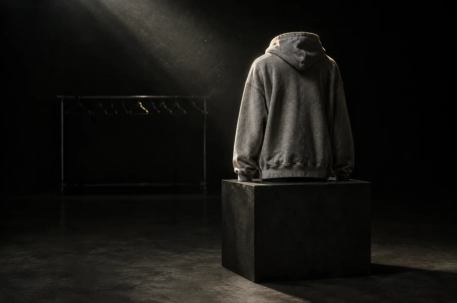 Ein verwaschener Kapuzenpullover steht allein auf einem dunklen Sockel im Spotlicht, dahinter eine leere Kleiderstange mit blanken Bügeln.