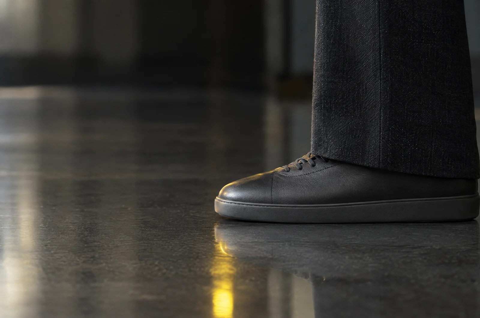 Ein flacher Ledersneaker steht dicht neben dem gerade fallenden Saum einer dunklen Stoffhose auf einem polierten Boden.
